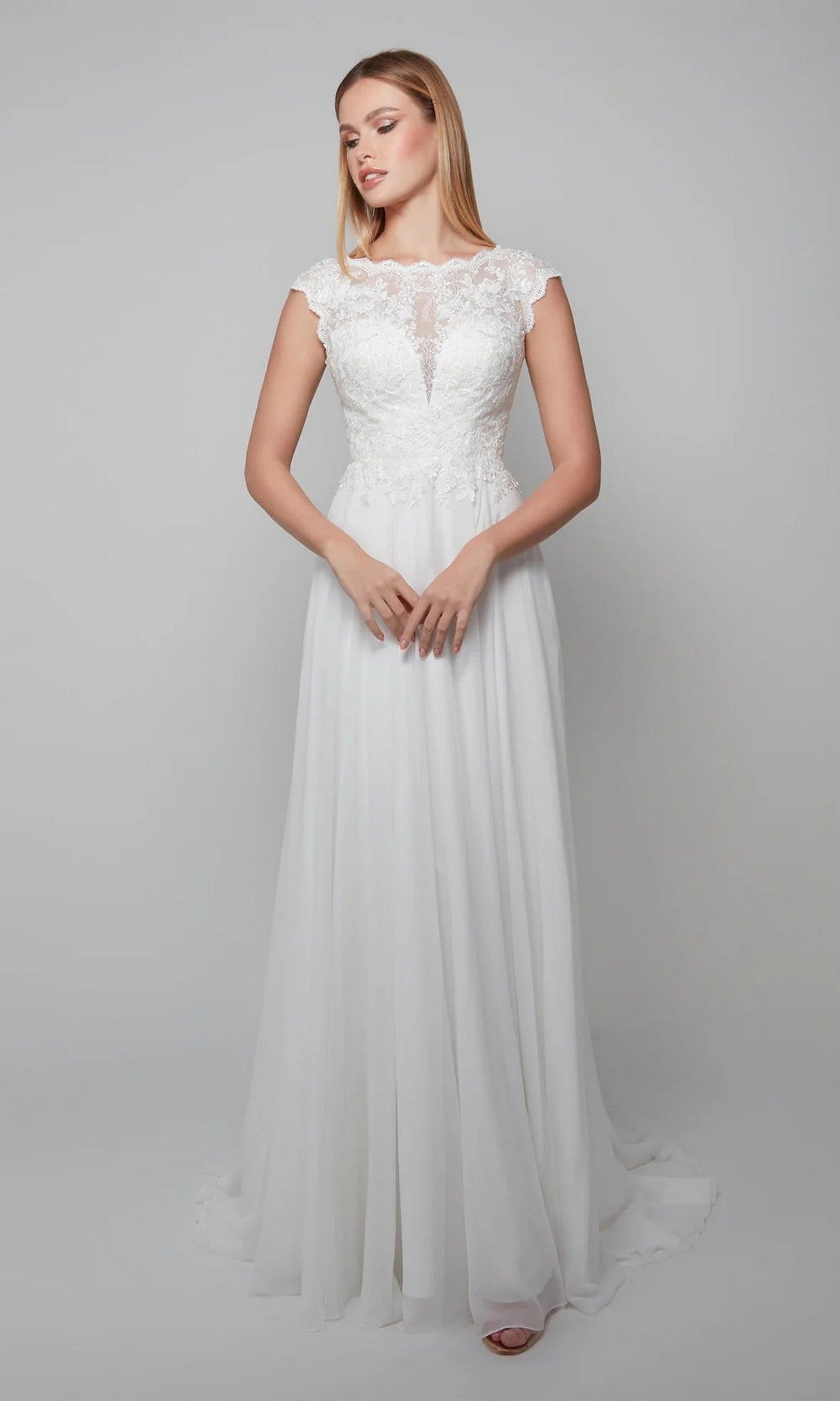 A woman in the Alyce 7073 long white dress stands against a plain light gray background, looking slightly to the side with her hands clasped in front of her.